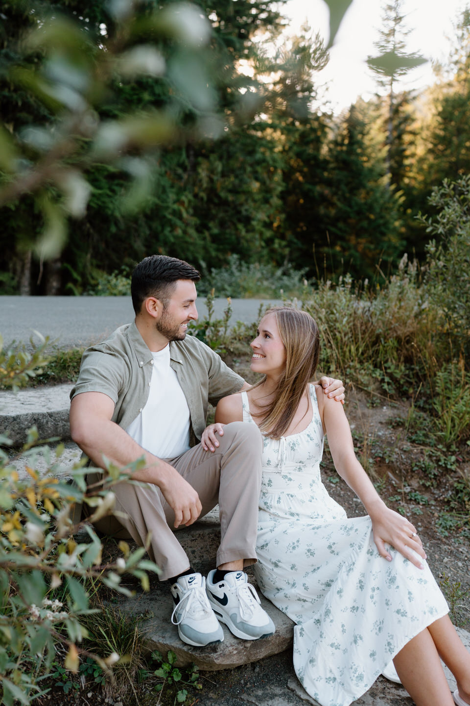 Couple sitting close and smiling softly at each other surrounded by greenery.