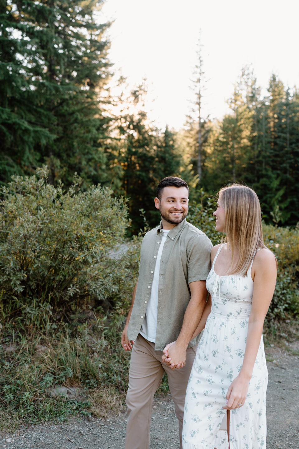 Couple smiling at each other while walking through greenery near the lake during a relaxed engagement session.