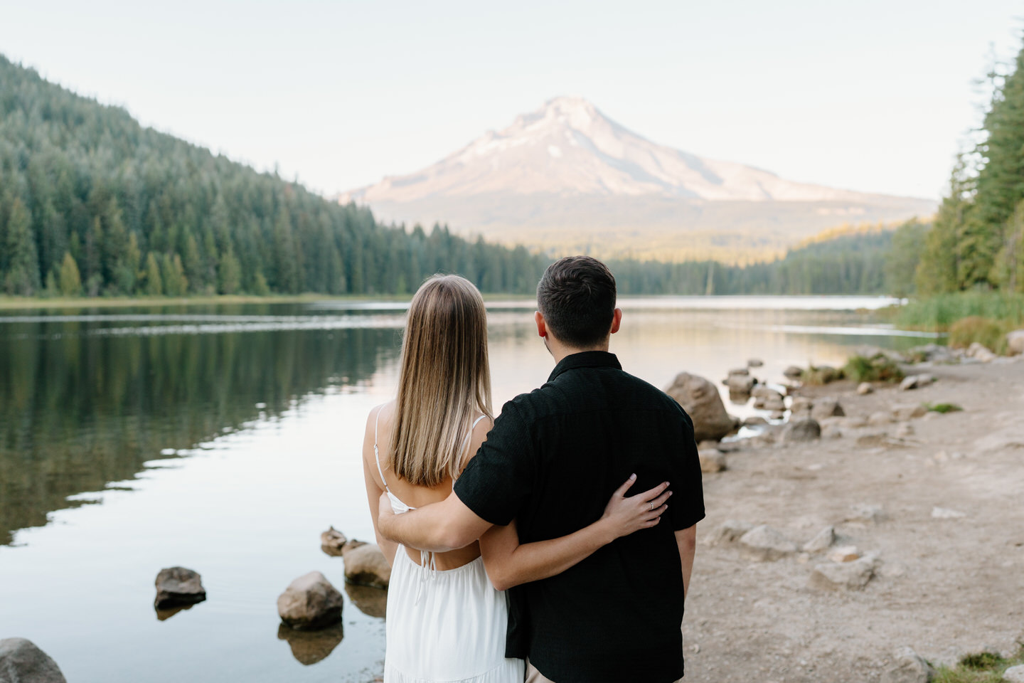 Back view of the couple embracing while looking across calm water and mountain views at Trillium Lake engagement session.