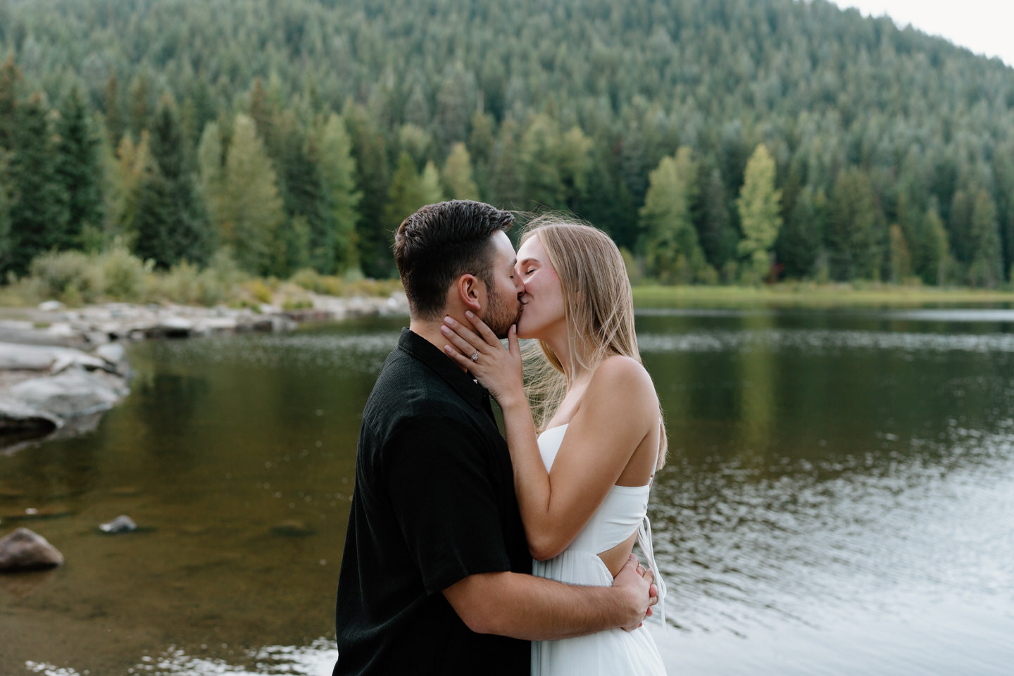 Romantic kiss by the water with evergreen trees surrounding the couple at the lake on a warm September evening.