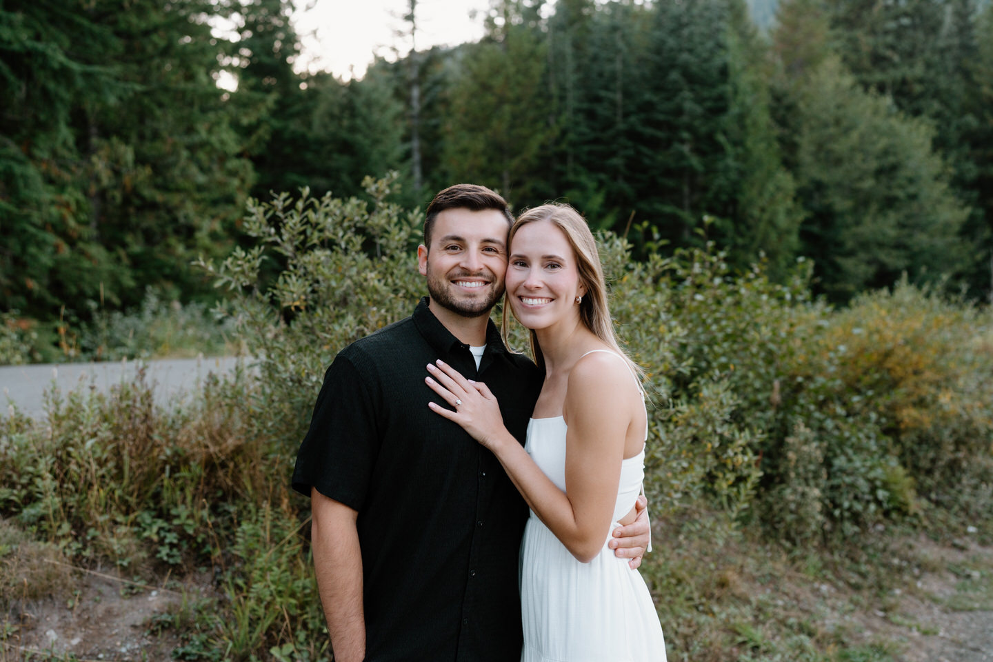 Couple smiling and holding each other in a forest clearing during engagement photos at the lake on a sunny September day.