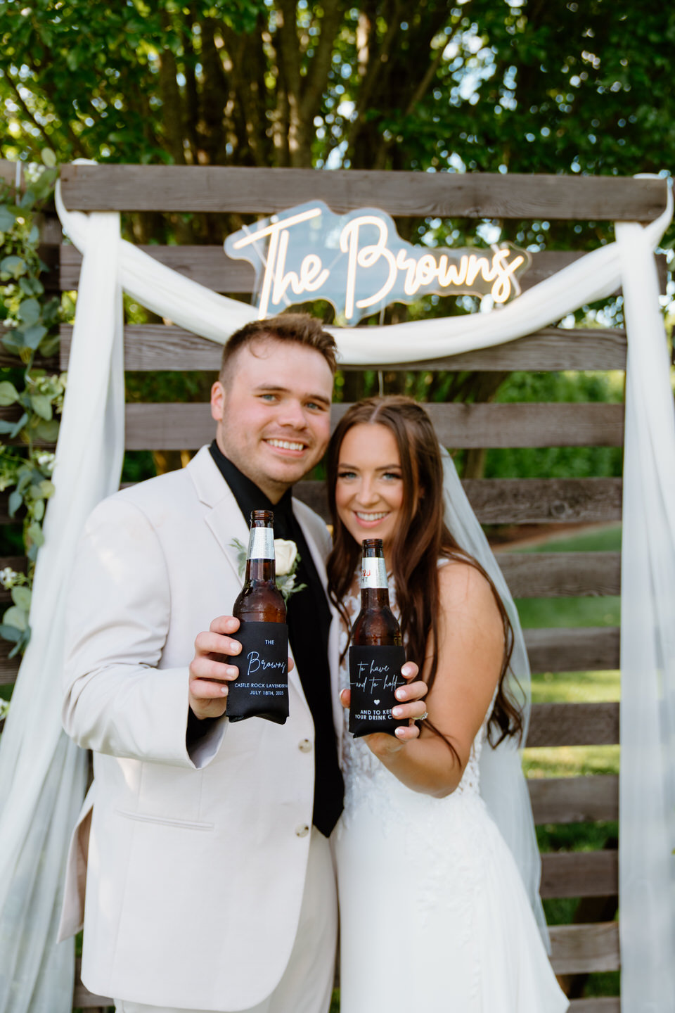 Hailey and Tyler holding custom beer koozies in front of a neon "The Browns" sign.