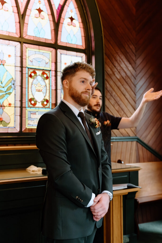 Groom listening with emotion as bride reads vows beneath stained glass windows at their Oaks Pioneer Church wedding ceremony.