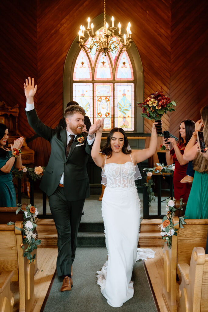 Newly married couple raise their hands in celebration as friends cheer inside the cozy chapel during an Oaks Pioneer Church wedding.