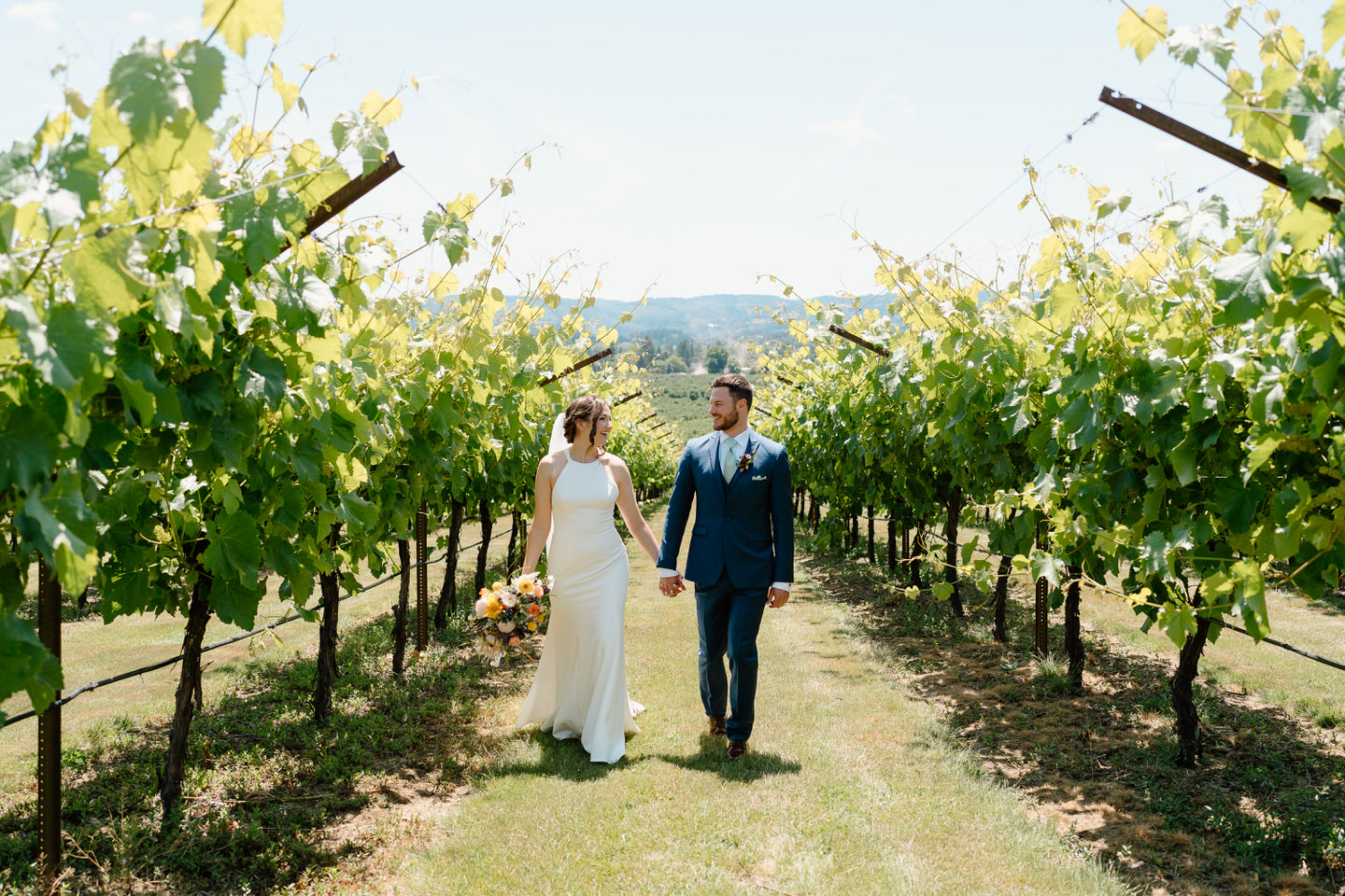 Bride and groom hold hands, looking cheerfully at each other as they walk through the vineyard at Scholls Valley Lodge.