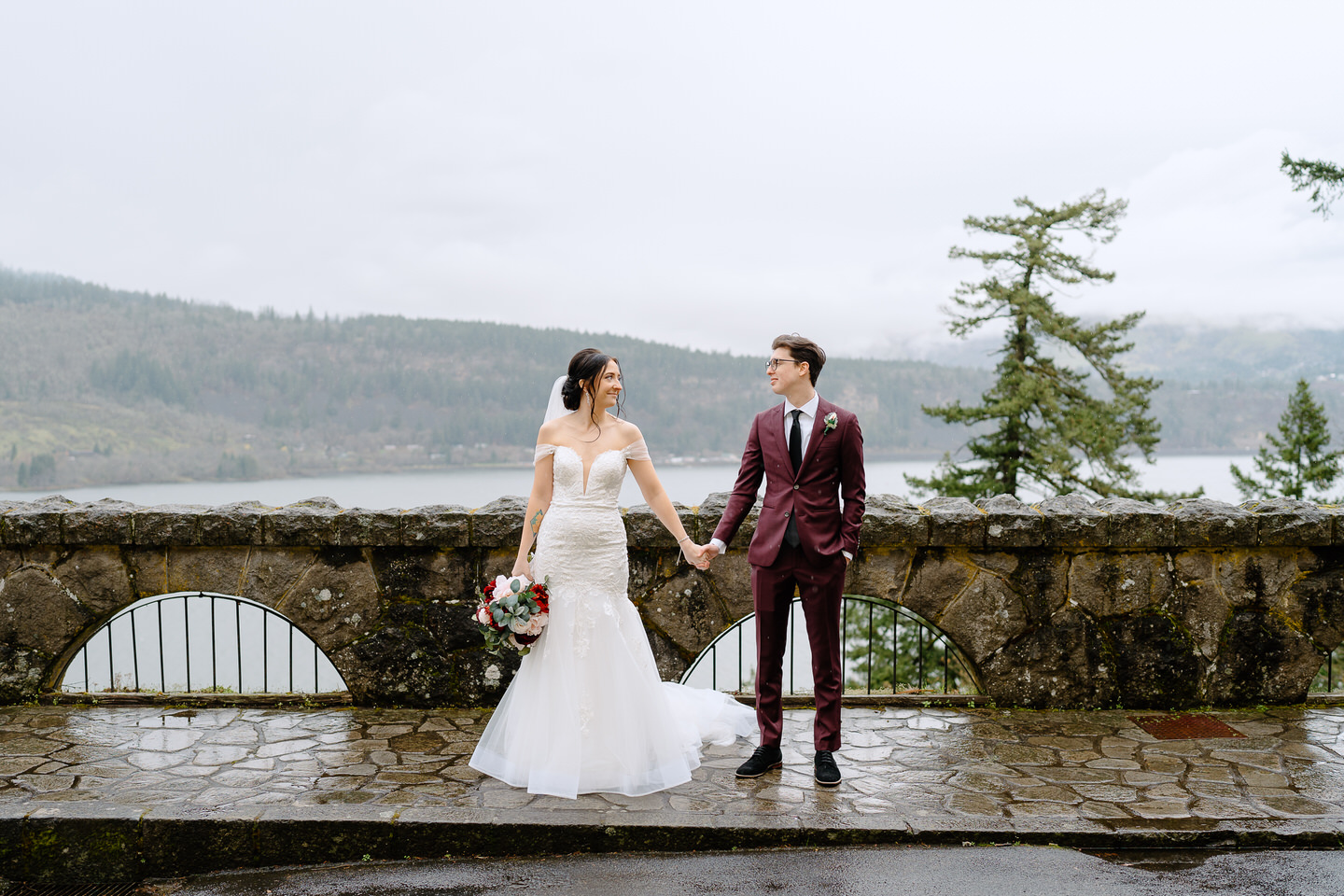 Couple holds hands and looks at each other with the rainy Columbia River Gorge behind them.