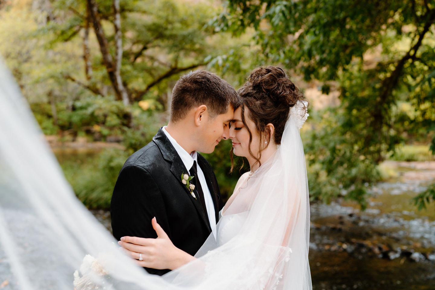 Bride and groom share a moment with her veil flowing beside her. The river is flowing behind them.