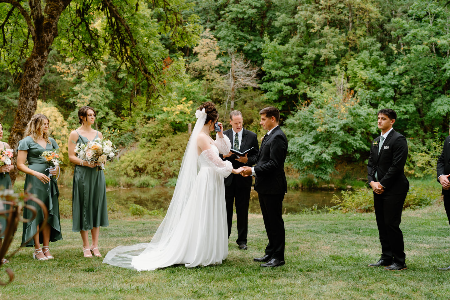 Bride and groom hold hands during their wedding ceremony. The river flows behind them in the garden.