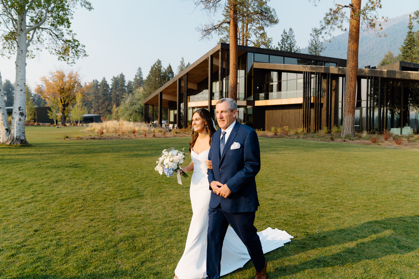 Bride walks down the aisle with her father at Black Butte Ranch, both smiling as guests watch against the stunning Central Oregon backdrop.