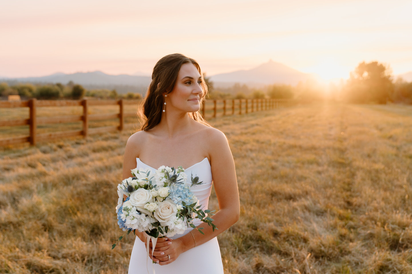 Bride standing at sunset, her gown glowing in the golden light, framed by the warm hues of the sky.