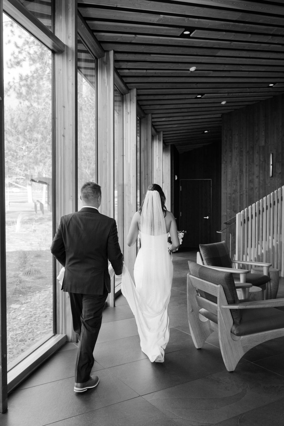 Black and white photo of the groom holding the bride's dress train as they walk through a building.