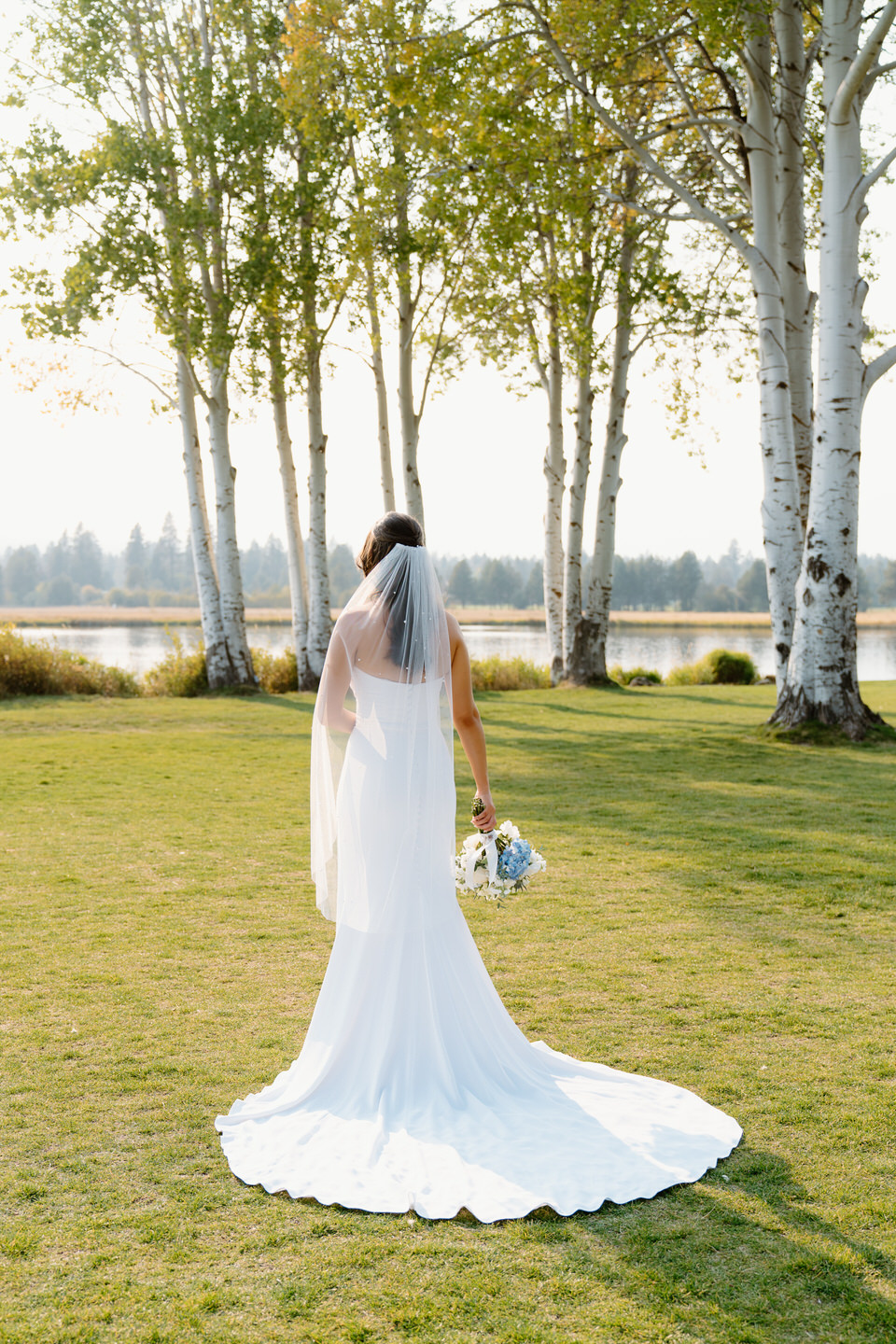 The bride stands gracefully, showcasing the intricate back of her dress and flowing veil against the scenic backdrop of Black Butte Ranch.