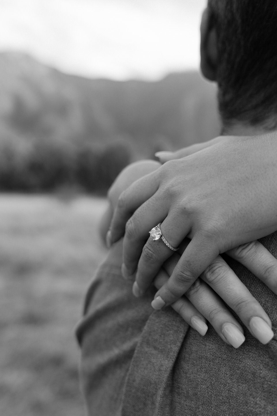 Hands wrapped around her fiance's back, showing off her round engagement ring.