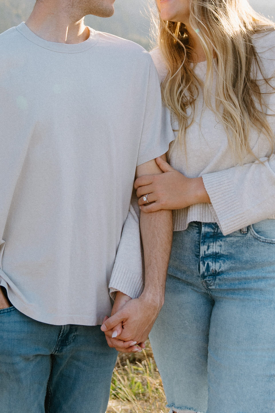 A close-up of intertwined arms with hands gently clasped, highlighting an engagement ring sparkling on the finger.