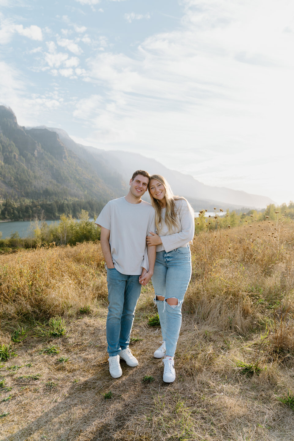 Engaged couple holding hands on a Columbia Gorge trail, with vibrant summer hues and river flowing in the background.