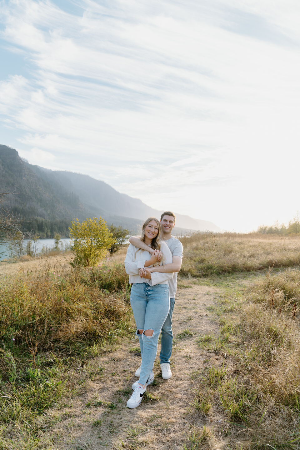 Couple with arms wrapped around each other, smiling warmly, framed by the Columbia Gorge's lush greenery and golden September light.