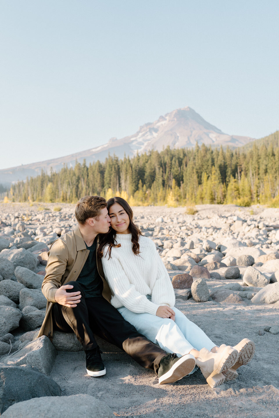 A couple sitting closely on a cluster of smooth gray rocks, with Mount Hood’s majestic snow-capped peak rising prominently in the background under a crisp autumn sky.