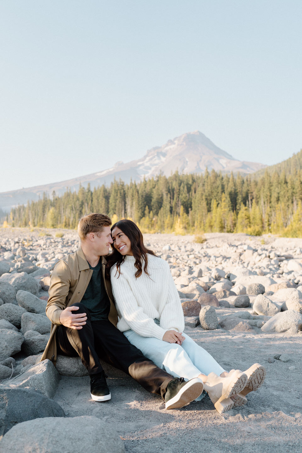 A candid moment of the couple laughing together while sitting on large boulders, the sweeping view of Mount Hood adding grandeur to the serene fall scene.