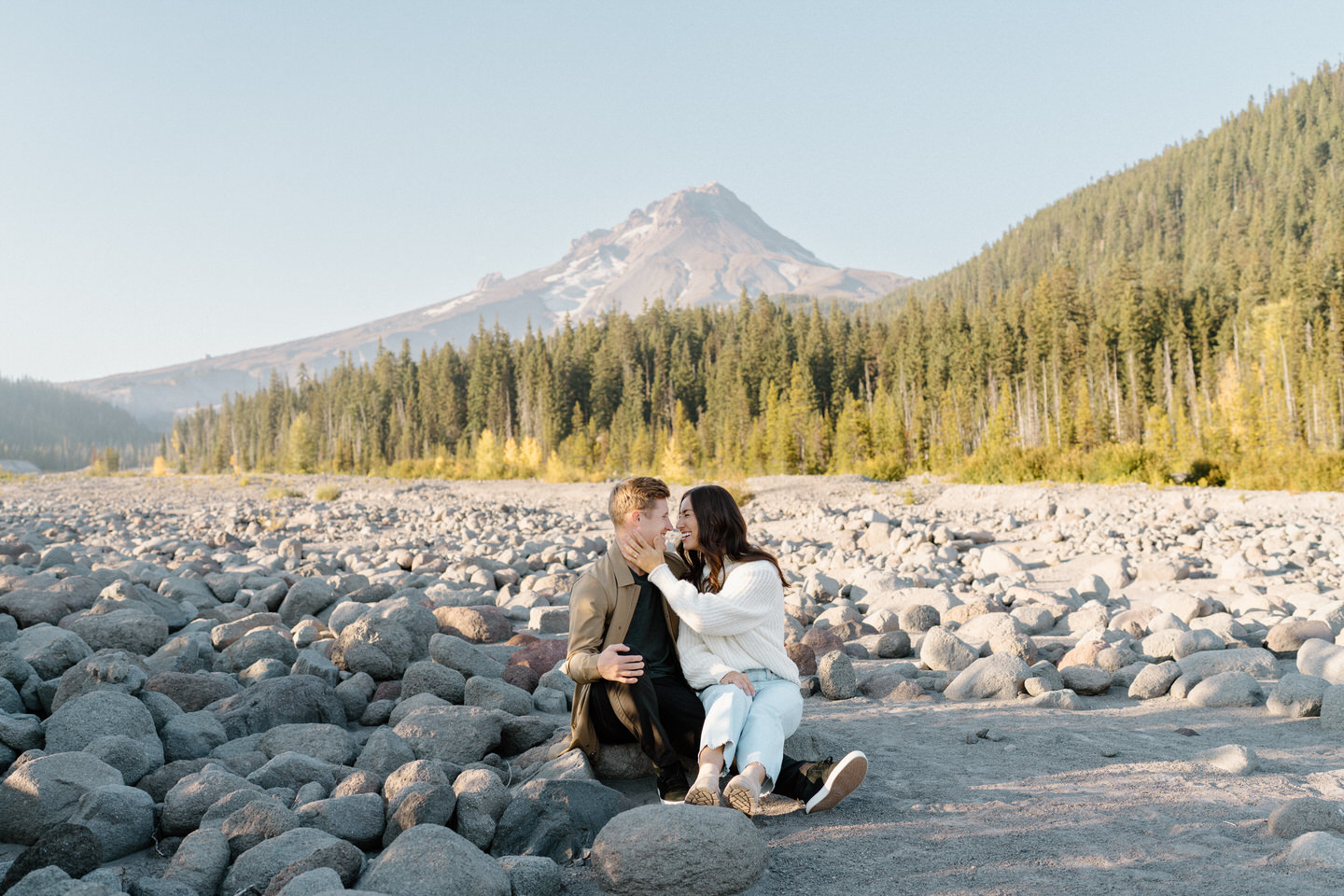 The couple sharing an intimate embrace as they sit on rocks overlooking the White River, with Mount Hood standing tall in the distance under soft golden light.