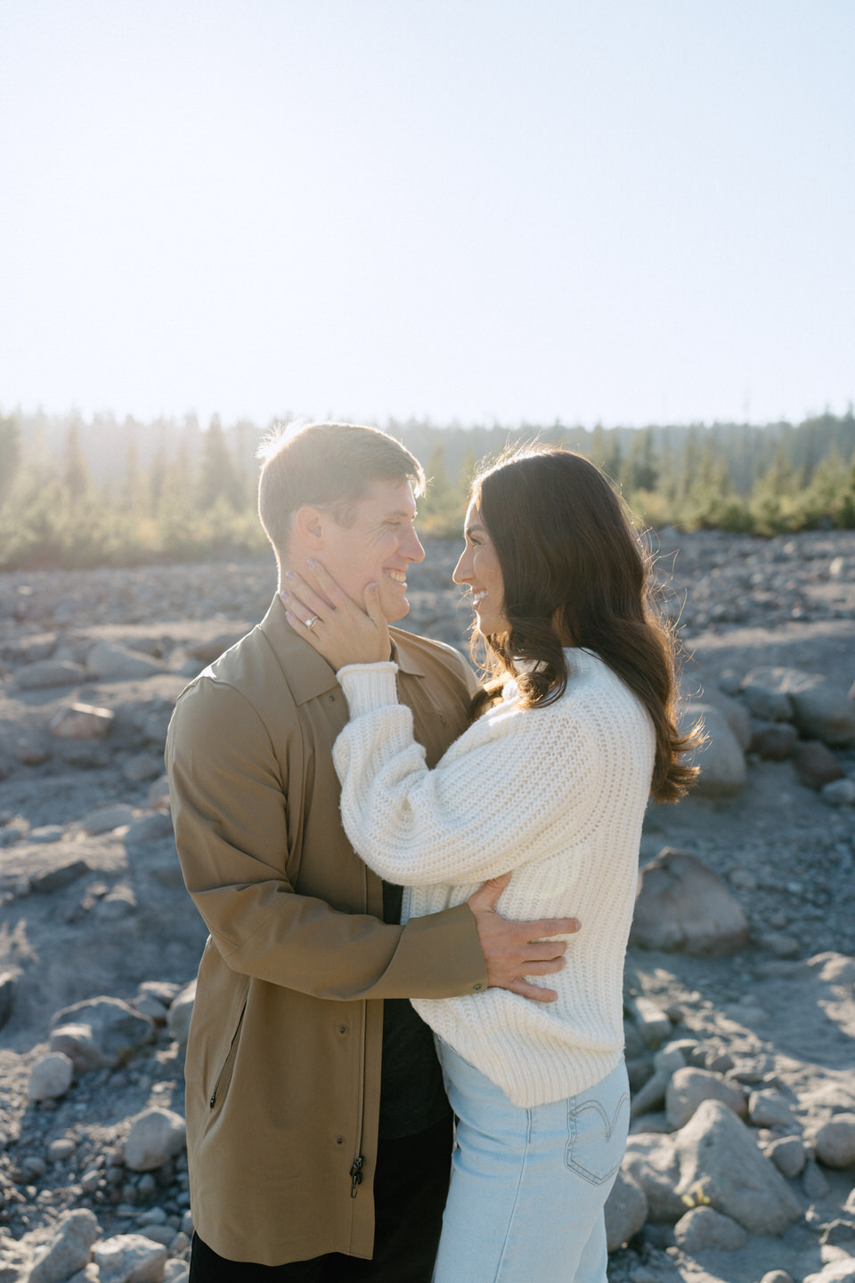 A couple laughing together while sitting on a large boulder by the White River, framed by the vibrant fall landscape and Mount Hood's snow-capped peak in the background.