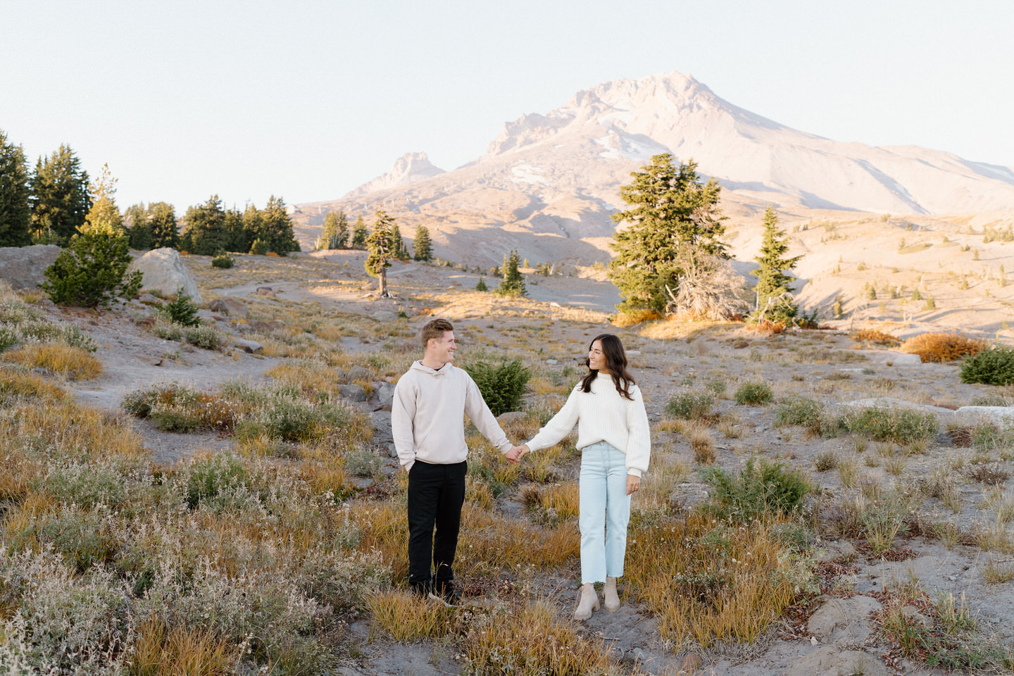 A couple holding hands and looking into each other's eyes with Mount Hood towering behind them in the sun.