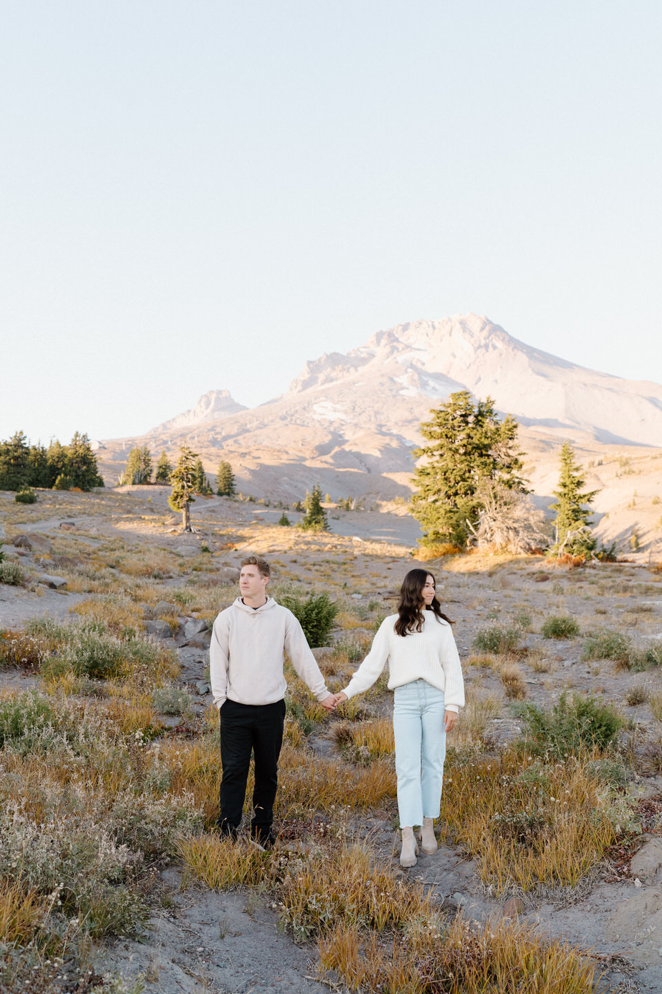 The couple holding hands on a trail, framed by golden autumn leaves and the towering peak of Mount Hood in the background.