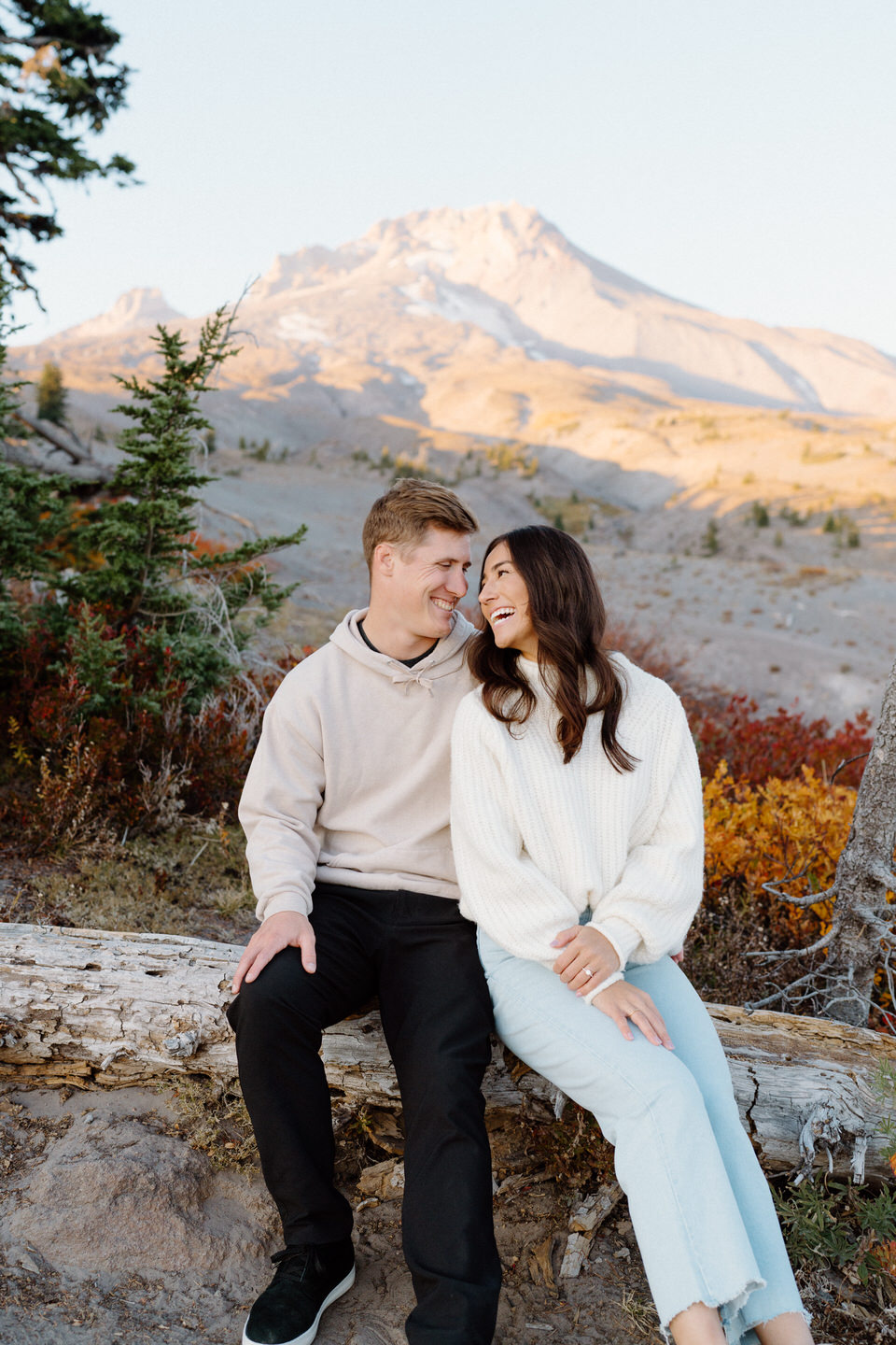 A couple sitting closely on a weathered log, surrounded by vibrant fall foliage, with Mount Hood’s snow-capped peak standing tall in the background.