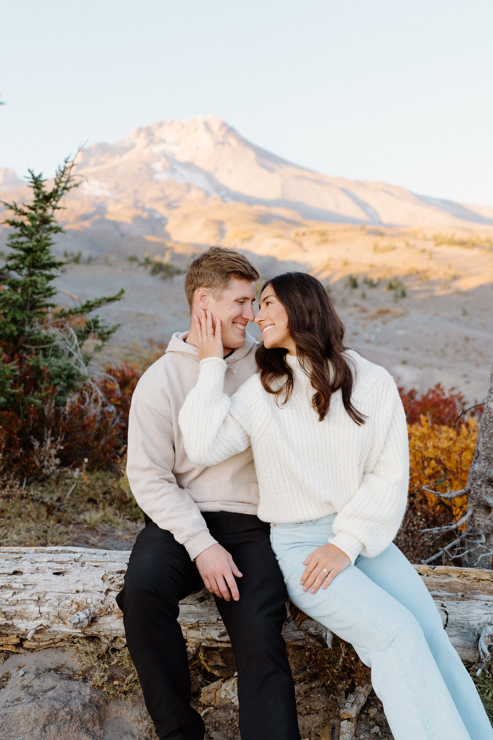 The woman holding her husband's face as they sit on a log, surrounded by rich fall colors, with Mount Hood towering serenely in the distance under soft afternoon light.