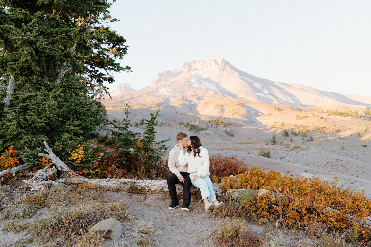 A candid shot of the couple laughing together on a fallen log, the warm autumn tones of the trees and the majestic Mount Hood creating a picturesque backdrop.