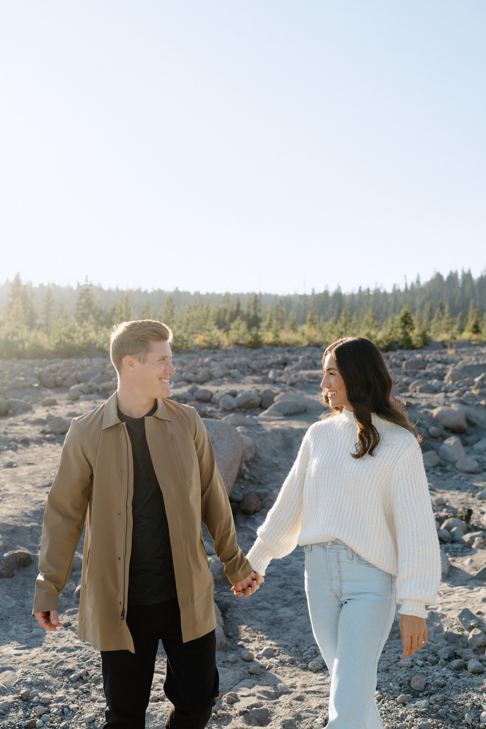 A man and woman walking hand in hand along a gravel path, their neutral-toned sweaters blending beautifully with the warm autumn colors at the base of Mount Hood.