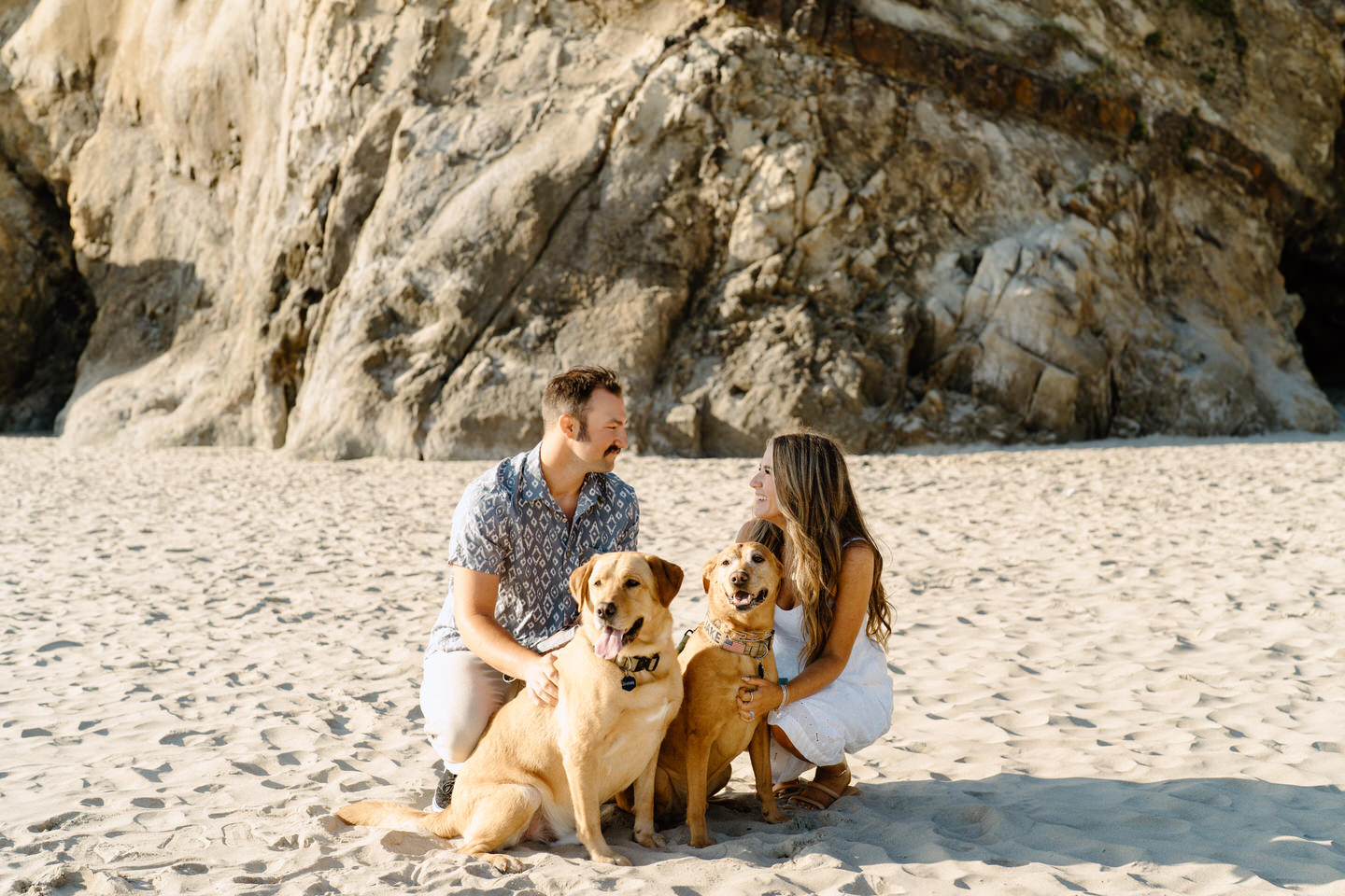 Couple with 2 yellow labs on the sand with rocks in the background